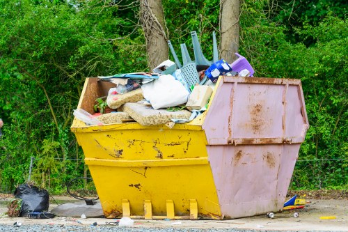 A large outdoor skip bin with a yellow lower section and a pink upper section, filled with assorted household rubbish including plastic bottles, foam, cardboard boxes, and gardening tools such as a watering can and hand trowels. The bin is positioned on a gravel surface adjacent to a background of dense green foliage and trees. Surrounding the bin are small scattered items and a black trash bag at the base, indicating a collection of garden clearance waste likely managed by a local rubbish removal service in Chelsea. The scene is well-lit by natural daylight, emphasizing the textures of the materials and the vibrant colours of the waste, in a setting that suggests ongoing garden or property clearance work typical of services provided by Garden Clearance Chelsea in the area.