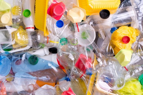 A large pile of discarded plastic bottles and containers, many with their caps still attached, in various colors including yellow, red, blue, green, and black. The bottles are made of clear or semi-transparent plastic, with some crumpled or flattened, and are scattered across what appears to be an outdoor or industrial setting, possibly a driveway or nearby area. Visible among the bottles are plastic packaging and caps of different sizes, with some of the bottles stacked or leaning against each other. The background hints at a concrete surface with no environmental features visible. This scene exemplifies typical rubbish that a waste removal service such as Garden Clearance Chelsea may collect and dispose of, especially in the context of garden or property clearance in the Chelsea area, post waste accumulation. The lighting is natural, highlighting the glossy textures of the plastic. The image emphasizes the importance of proper rubbish collection and disposal of plastic waste.