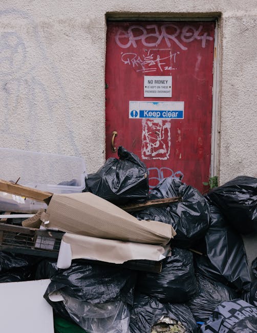 A collection of black plastic rubbish bags and discarded cardboard and packaging materials are piled in front of a red metal door set into a beige, textured concrete wall. The door has various graffit