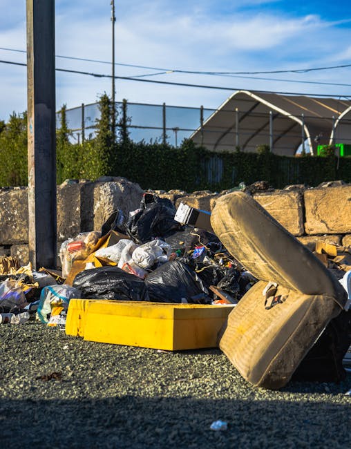 A narrow urban alleyway cluttered with a large, grey, weathered garbage bag in the foreground, positioned on a concrete surface stained with dirt and debris. Behind it, a mountain of mixed waste mater
