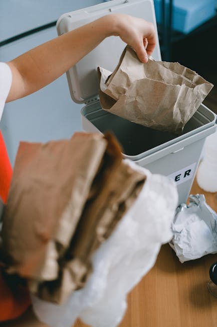 A person disposing of paper waste by placing crumpled brown paper bags into a grey rubbish bin, positioned on a wooden surface indoors. The person's hand is visible, gripping a paper bag as it is bein