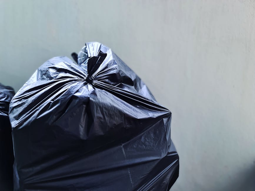 A person disposing of paper waste by placing crumpled brown paper bags into a grey rubbish bin, positioned on a wooden surface indoors. The person's hand is visible, gripping a paper bag as it is bein